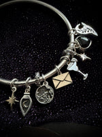 A silver charm bracelet with multiple whimsical charms, including a small owl, a star, a potion bottle, a coin, an envelope, and a martini glass, displayed on a dark textured background.