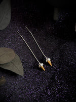 A pair of long silver-toned drop earrings with small cone-shaped amber or honey-colored pendants, displayed on a dark textured fabric with decorative leaves.