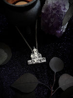 A silver-toned pendant necklace featuring an engraved house or cottage design on a delicate chain, displayed on a dark textured surface with decorative leaves, a black bowl, and a purple crystal cluster.